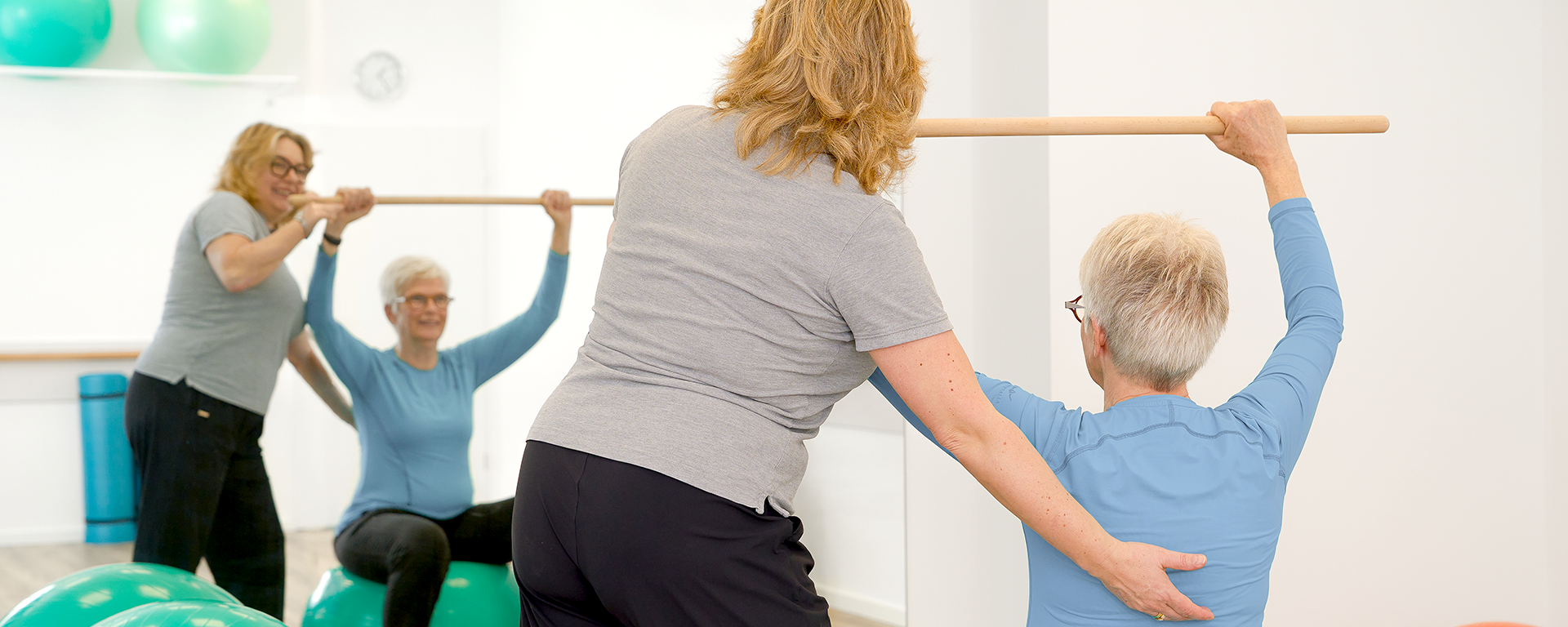 Eine Patientin sitzt auf dem Sitzball und hält einen Stab in die Höhe. Die Therapeutin unterstützt die Bewegung.
