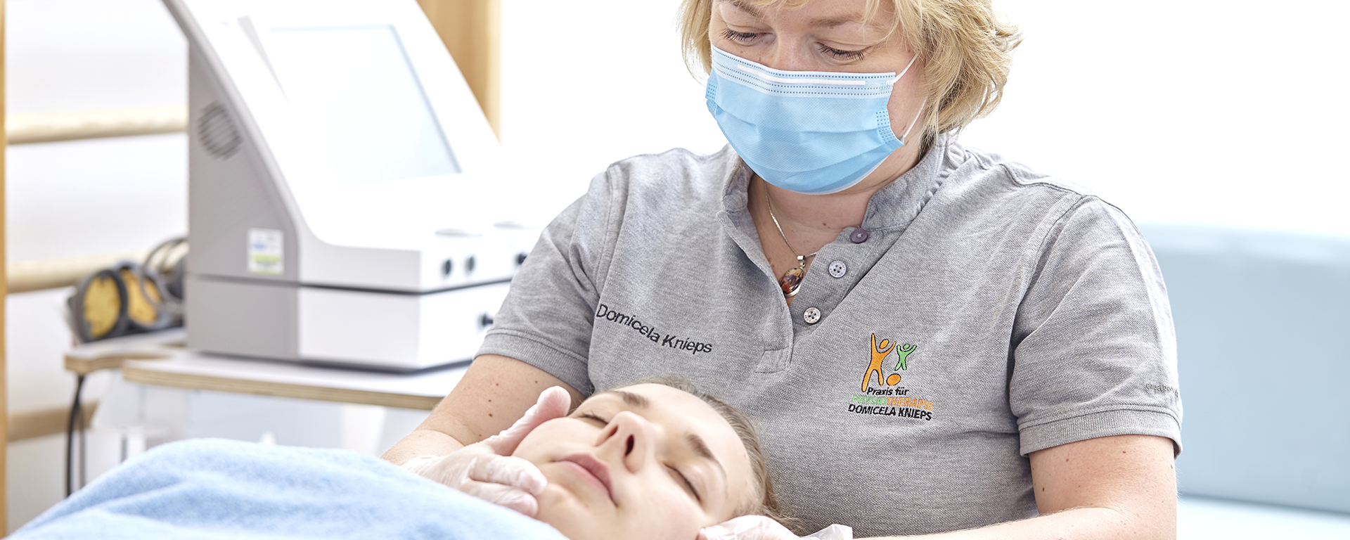 Die Therapeutin hat eine Maske an und beugt sich zu der liegenden Patientin um deren Kiefer von außen abzutasten.