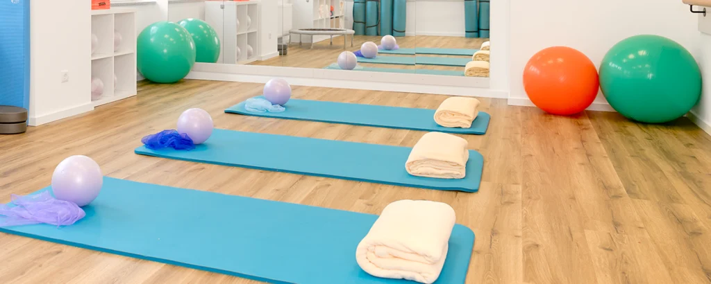 Drei Gymnastikmatten mit Handtüchern und Bällen liegen auf einem Holzboden vor einem großen Spiegel.