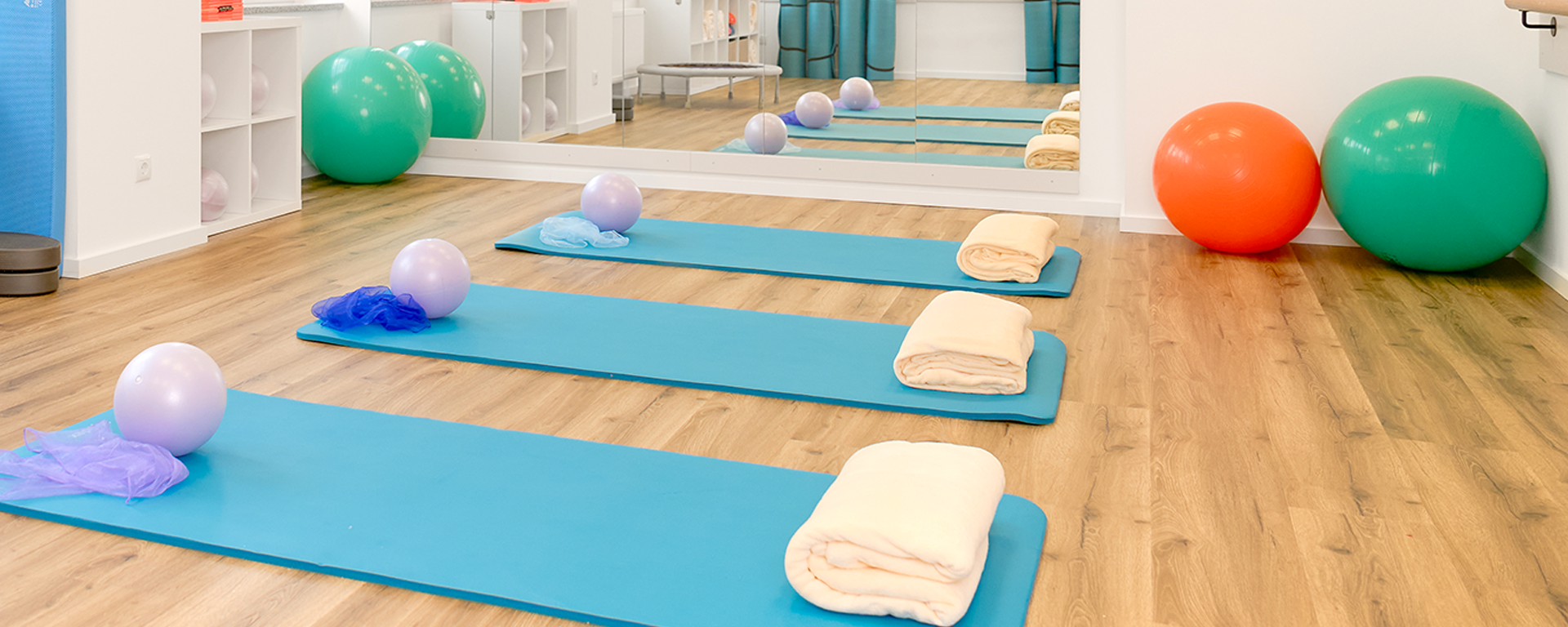 Drei Gymnastikmatten mit Handtüchern und Bällen liegen auf einem Holzboden vor einem großen Spiegel.