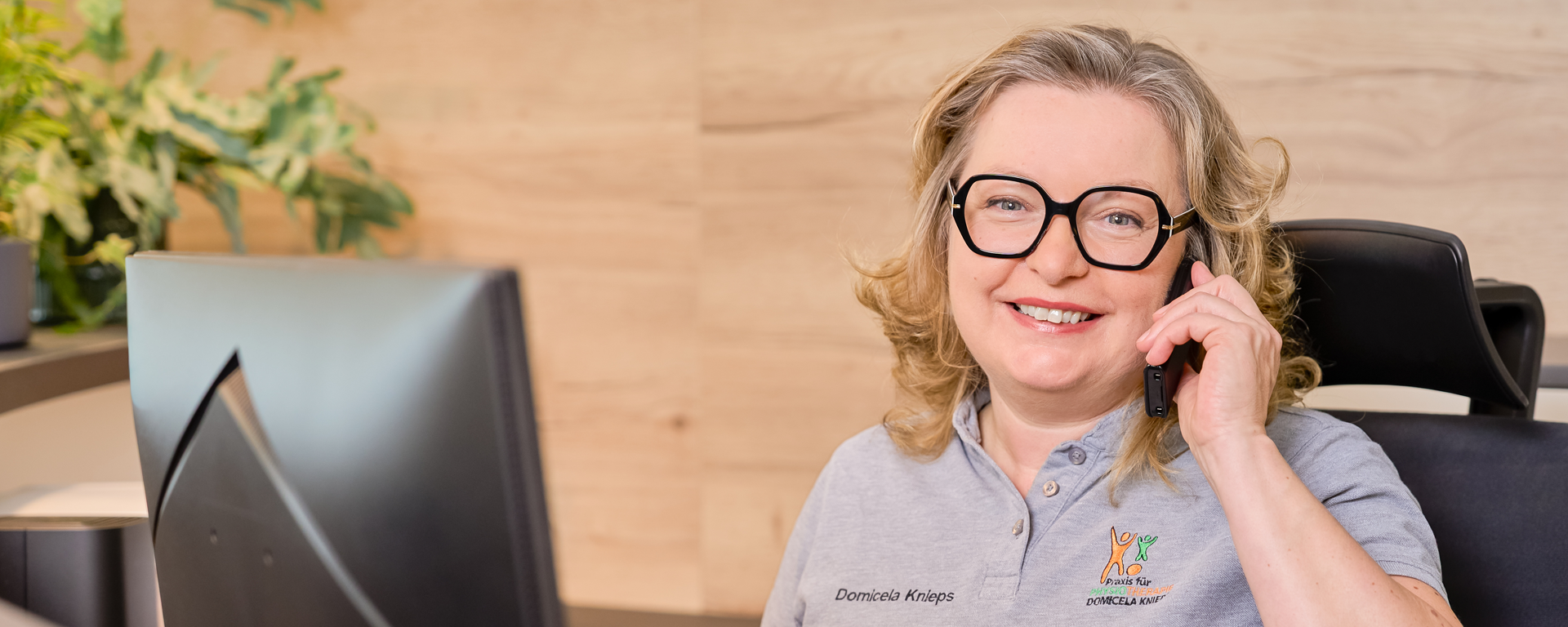 Domicela Knieps sitzt an der Rezeption, schaut freundlich in die Kamera und telefoniert.